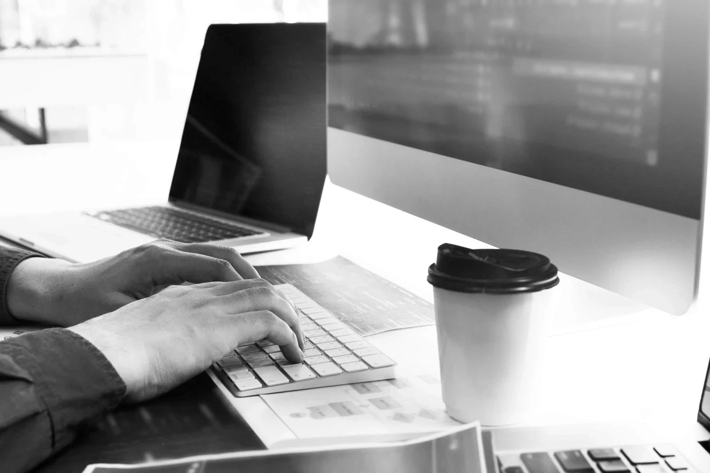 Schwarz-weißes Foto einer Person beim Arbeiten an einer Tastatur vor einem Computerbildschirm mit Kaffeebecher auf dem Schreibtisch.